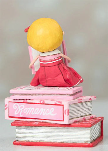 Hand-painted “Love Story” figurine by Lori Mitchell—sweet girl in pink and red dress perched atop a stack of romance books, lost in a love story